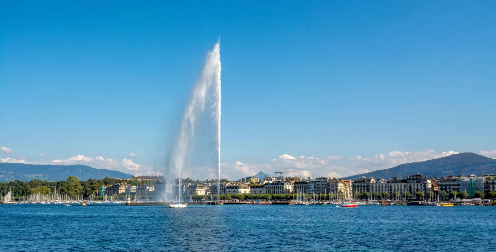 Vue panoramique du Jet d’eau de Genève sur le lac Léman, avec les immeubles en arrière-plan et les montagnes à l’horizon