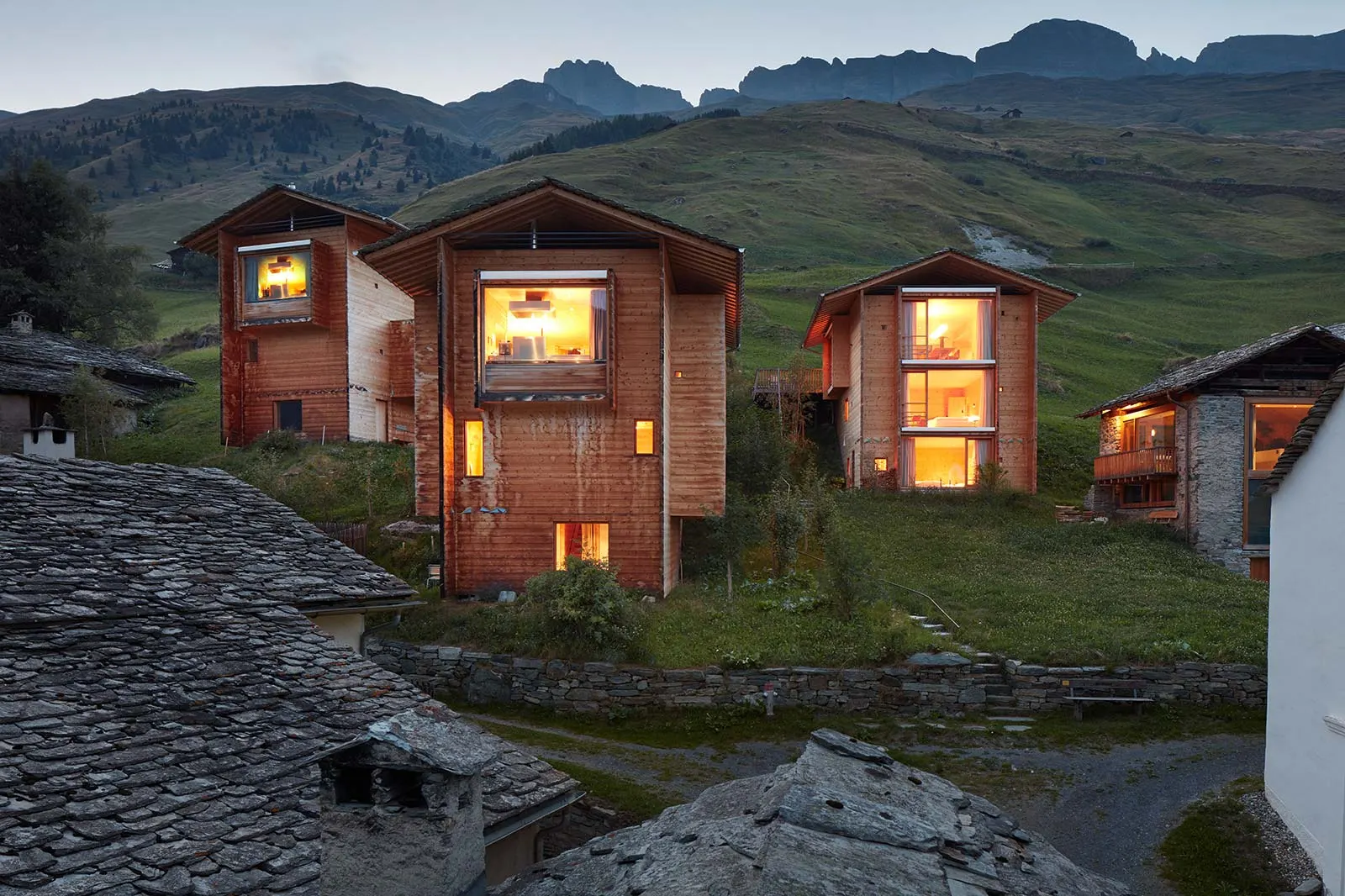 Les trois maisons en bois conçues par Peter Zumthor dans le hameau de Leis, près de Vals, éclairées à la tombée du jour dans le paysage de montagne